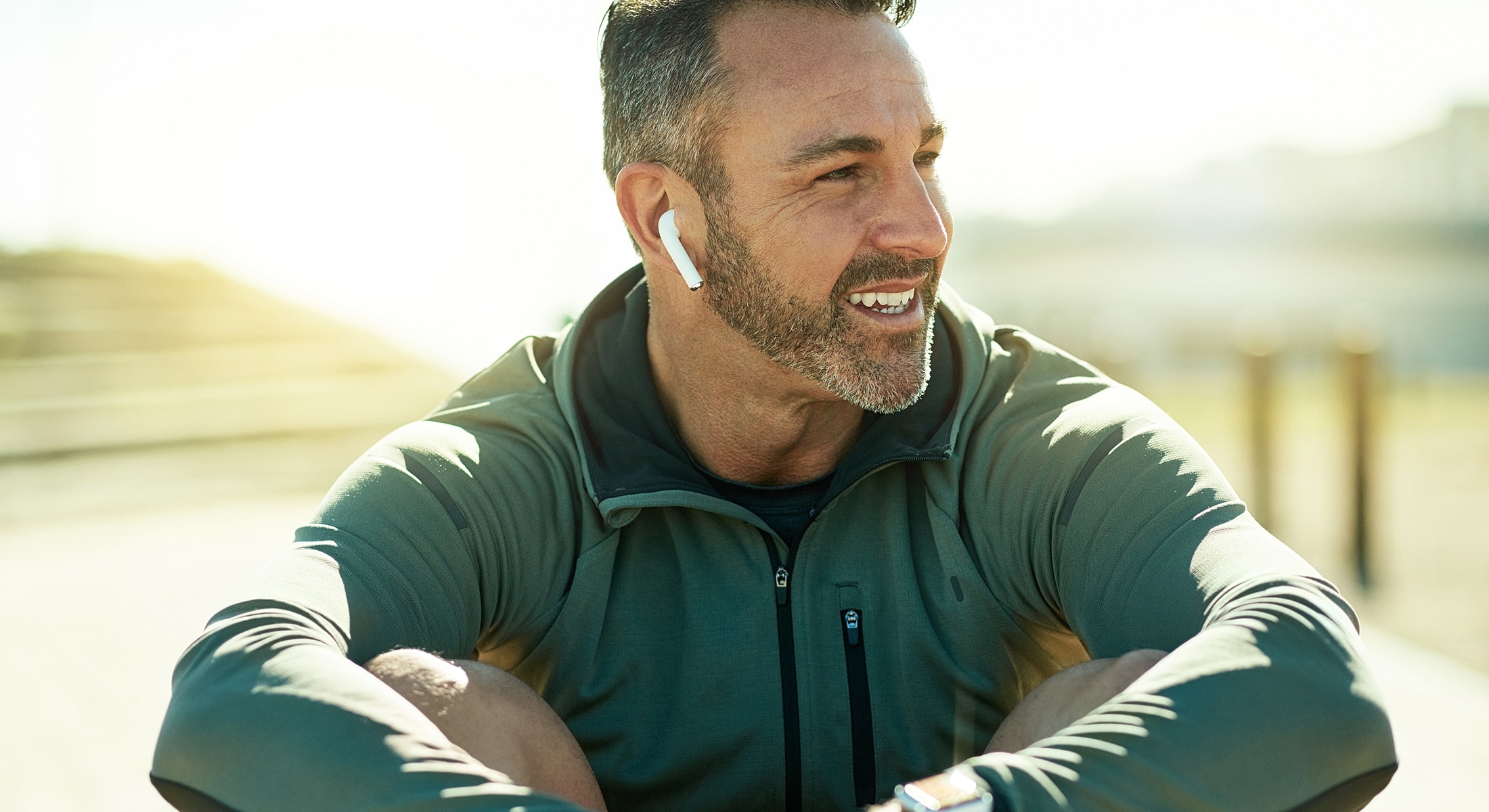 Man smiling with earbuds outdoors, sunlight background.